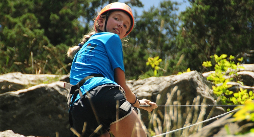 A young person wearing safety gear is secured by ropes as they look down toward the camera while rock climbing. 